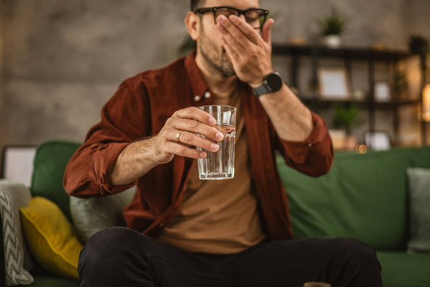 Дорослий білий чоловік сидить на дивані і п'є медичні таблетки вдома - Фото, зображення