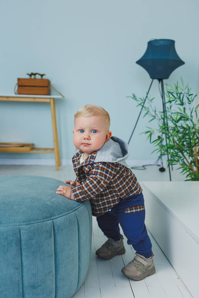 A 9-month-old baby boy in a stylish shirt, pants and sneakers is standing near a support. - Photo, image