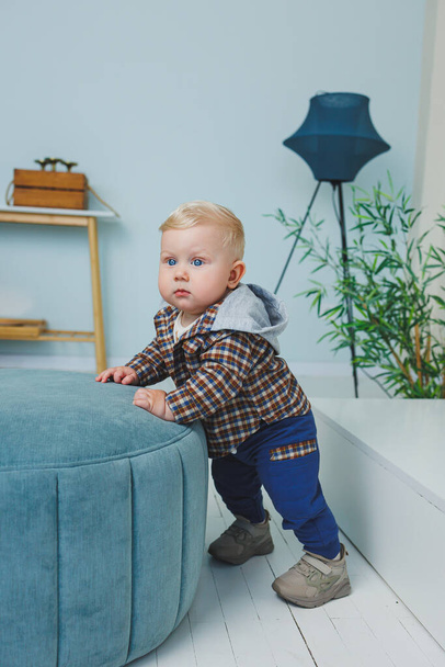 A 9-month-old baby boy in a stylish shirt, pants and sneakers is standing near a support. - Photo, image