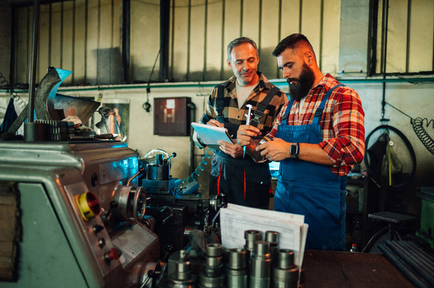 A machinist consulting with a senior engineer about a metal piece in a factory. The engineer is holding a clipboard, comparing measures. The metallurgist is holding a caliper. Factory setting with various machines and pieces. - Fotoğraf, Görsel