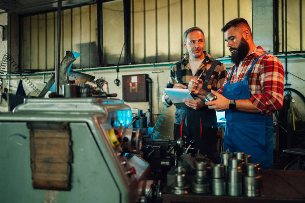 Two metal workers checking if metal pieces are correct measure. Senior man is holding a clipboard, comparing numbers. Young man with a beard is holding a machine piece and a caliper. Overalls, shirts. - Fotoğraf, Görsel