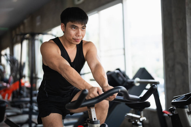 Ένας αποφασισμένος άνδρας με μαύρο μπλουζάκι εκτελεί ποδηλατική προπόνηση σε σταθερό ποδήλατο σε σύγχρονο γυμναστήριο. Η συγκεντρωμένη έκφραση και η μυϊκή του δομή τονίζουν την αφοσίωσή του στη φυσική κατάσταση. - Φωτογραφία, εικόνα
