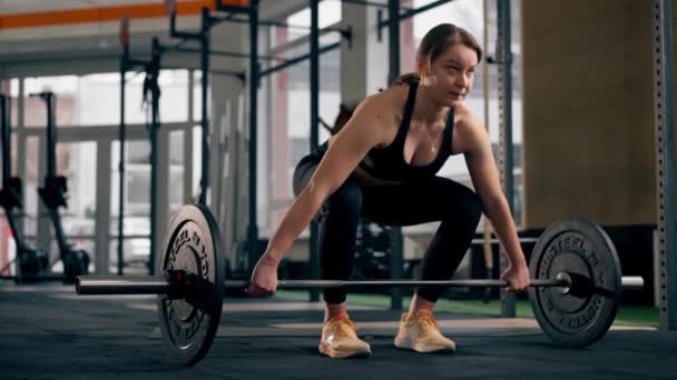 en el gimnasio una chica entrenador en un top negro está de pie haciendo ejercicios con una barra y pesas - Metraje, vídeo