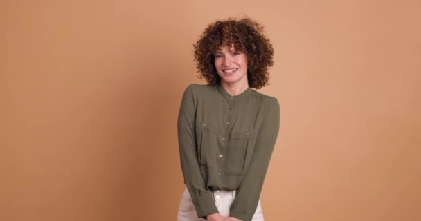 Laughing young curly haired female in makeup and full sleeves round neck top looking at camera while posing with moving head and standing on beige background - Footage, Video