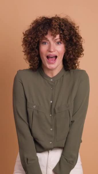 astonished young curly haired female in makeup and opened mouth looking at camera while standing with folded hands and touching hair on beige background. Vertical orientation - Footage, Video