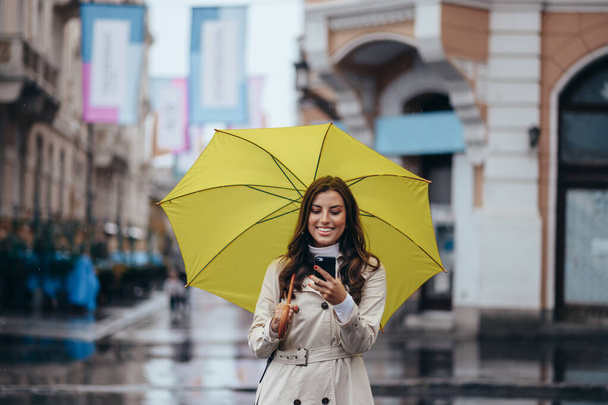 Schöne junge Frau mit Smartphone und gelbem Regenschirm an einem regnerischen Tag in der Stadt - Foto, Bild