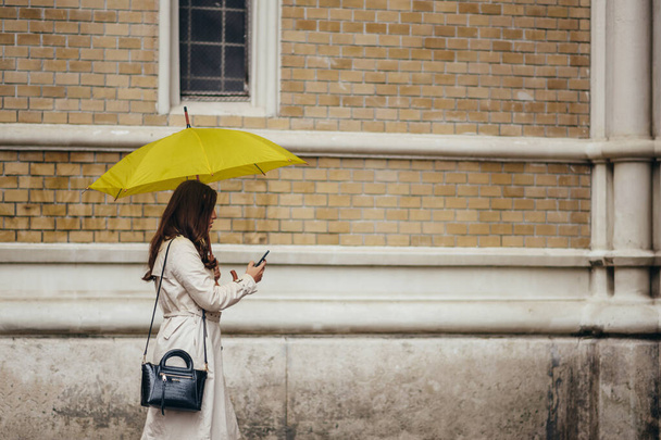 Schöne junge Frau mit Smartphone und gelbem Regenschirm an einem regnerischen Tag in der Stadt - Foto, Bild