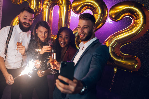 Happy group of friends celebrate New Year's Eve while drinking champagne on a party in a club and taking selfie with a smartphone - Фото, изображение