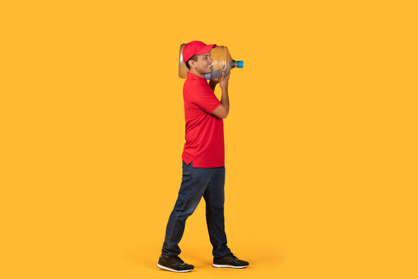 Drlivery άνθρωπος σε ένα κόκκινο πουκάμισο και καπάκι μεταφέρουν ένα μεγάλο γαλόνι νερό πάνω από τον ώμο του, ενώ στέκεται πάνω από ένα στερεό φωτεινό κίτρινο φόντο. - Φωτογραφία, εικόνα