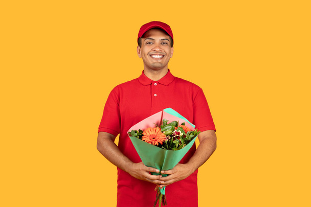 Kırmızı üniformalı neşeli bir teslimatçı canlı bir buket çiçek tutuyor, mutluluk saçıyor. Sarı zemin neşeli atmosferi vurguluyor.. - Fotoğraf, Görsel