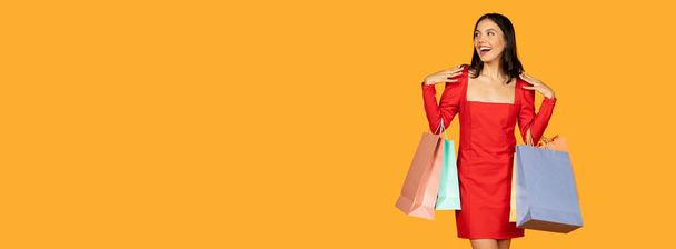 Молодая женщина с радостным выражением лица носит с собой несколько сумок для покупок, одетая в стильное красное платье. Она, кажется, на полпути, возможно, идет, панорама с копировальным пространством - Фото, изображение