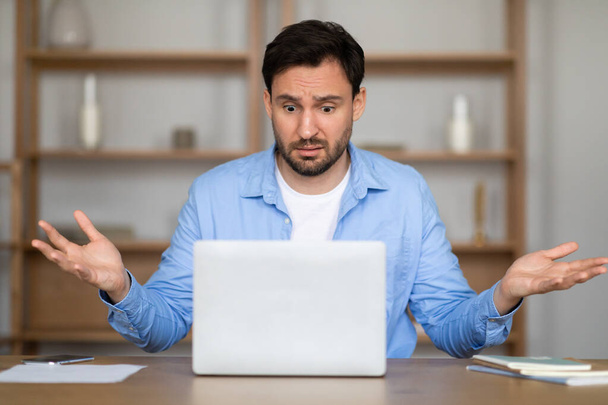 カジュアルなブルーのシャツを着た男性が机に座り,ノートパソコンのスクリーンを見ていると不確実に腕を上げて,困惑する仕事の状況に対処する. - 写真・画像