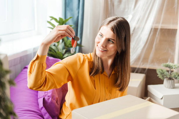 Smiling beautiful woman holding keys to new apartment, sitting near packed cardboard boxes in living room. Concept of rent, real estate - Foto, Imagem