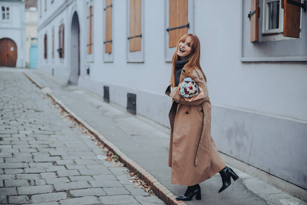 ファッショナブルなレトロなレトロなレッドヘッドの女性の屋外ファッション写真は,彼女の手の花とカメラで笑顔で街路を歩いています. 市街地の花束を持つ若い女性の完全な長さ. - 写真・画像