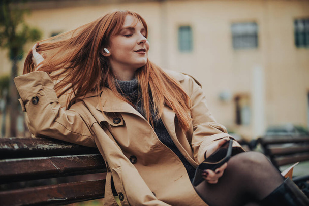 Egy divatos vörös hajú nő retro ruhában ül egy padon a város utcáján fülhallgatóval és élvezi a zenét. Portré egy bájos hölgyről, aki az utcán ül telefonnal a kezében. Fénymásolási hely. - Fotó, kép