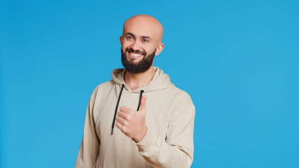 Arab férfi modell ad egy hüvelykujj fel a stúdióban, kifejezve elégedettségét a kék háttér. Közel-keleti fickó, aki jóváhagyást és gesztust mutat, pozitív szimbólum. Kettes kamera. Kézi lövés.. - Fotó, kép