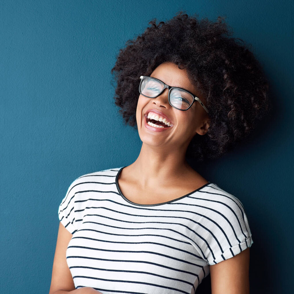 Portrait, funny and woman with glasses, afro and confident girl on blue studio background. Face, person and model with eyewear, cheerful and laughing for joke, humor and happiness with smile or gen z. - Фото, изображение