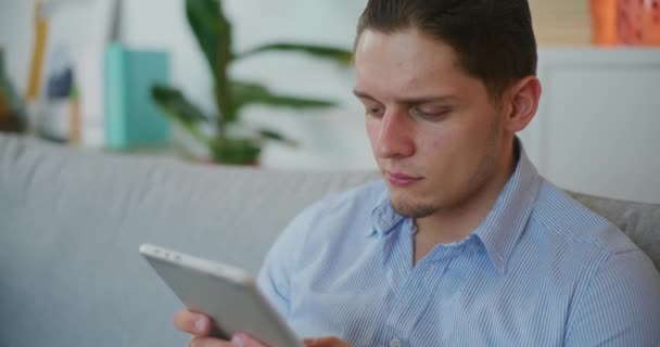 Confident man uses digital tablet sitting on sofa browsing social media - Footage, Video