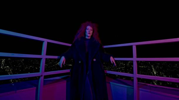 Fashion woman on roof of high-rise. Media. Stylish woman in photo shoot on roof of high-rise at night. Neon photo shoot with woman in coat on high-rise. - Photo, Image