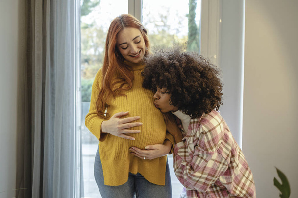 Těhotná žena ve žlutém svetru s úsměvem, jak její přítel políbí její břicho oknem. - Fotografie, Obrázek