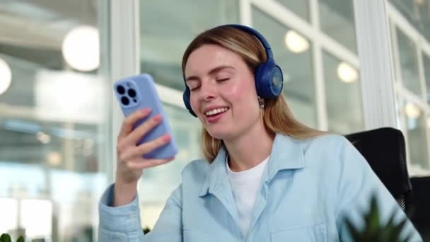 Aktive kaukasische Geschäftsfrau genießt energiegeladene Musik in drahtlosen Kopfhörern, während sie ihr persönliches Smartphone in der Hand hält. Hübsche Dame sitzt am persönlichen Arbeitsplatz mit digitalem Gerät drinnen. - Filmmaterial, Video
