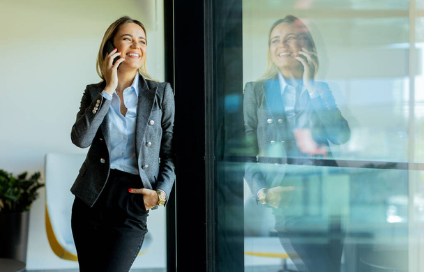 Una donna professionale sorride mentre parla al telefono in un ufficio luminoso e moderno. La luce del sole filtra attraverso grandi finestre, creando un'atmosfera calda - Foto, immagini