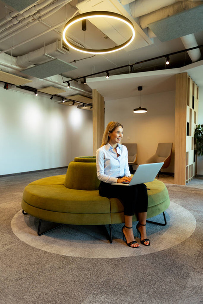 Woman seated on a green circular couch types on her laptop in a modern office lounge area under sleek, contemporary lighting - Photo, Image