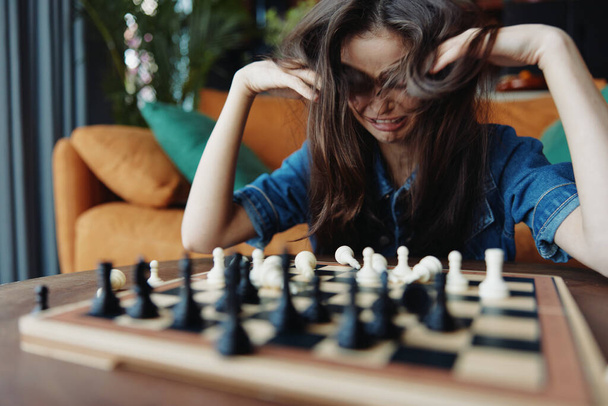 Mujer estresada jugando al ajedrez en la mesa frente al sofá con la cabeza en las manos - Foto, Imagen