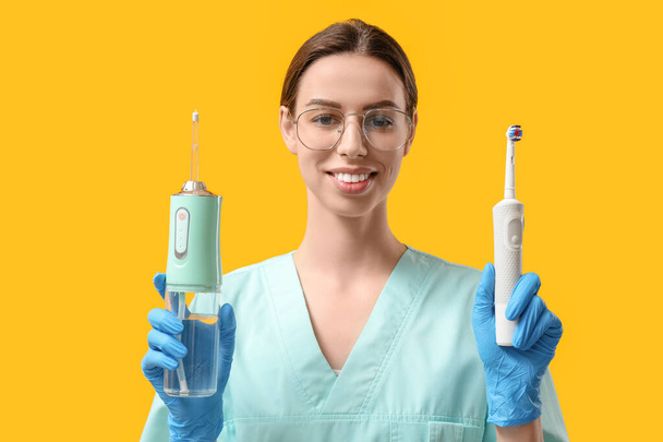 Женщина-стоматолог с оральным поливом и электрической зубной щеткой на желтом фоне - Фото, изображение