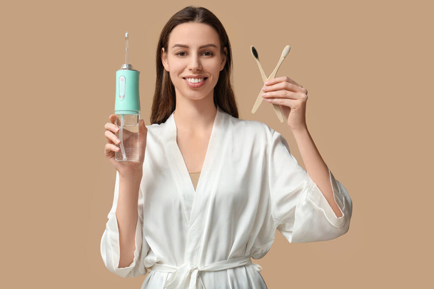 Krásná mladá žena s orální zavlažovač a zubní kartáčky na hnědém pozadí - Fotografie, Obrázek