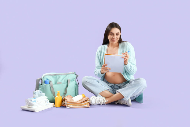 Mujer embarazada joven con bolsa de hospital de maternidad escritura lista de verificación sobre fondo lila - Foto, imagen