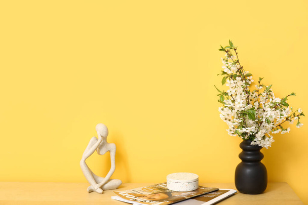 ramos florescentes em vaso, estatuetas e revistas na mesa perto da parede laranja - Foto, Imagem