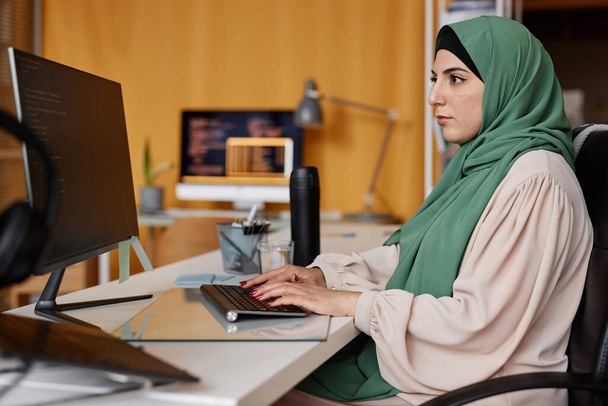 Portrait latéral de la femme musulmane réussie en tant que programmeuse féminine écrivant le code au bureau et portant des vêtements modestes - Photo, image