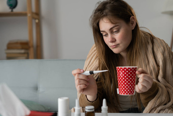 Eine kranke Frau auf dem Sofa blickt auf das Thermometer und trinkt Tee. Fiebersenkend gegen Fieber. Virusinfektion. - Foto, Bild