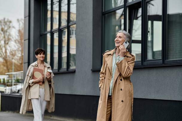 Two women, mature beautiful lesbian couple, walk down the street talking on their cell phones. - Photo, Image