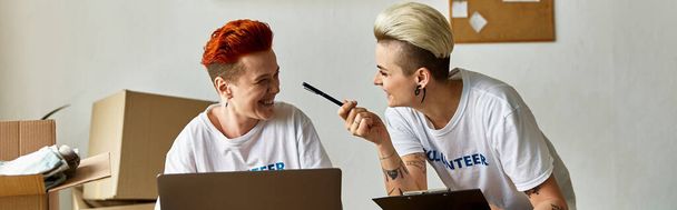 Молодая пара лесбиянок, одетых в футболки добровольцев, глубоко сосредоточена, работая над своим ноутбуком вместе. - Фото, изображение