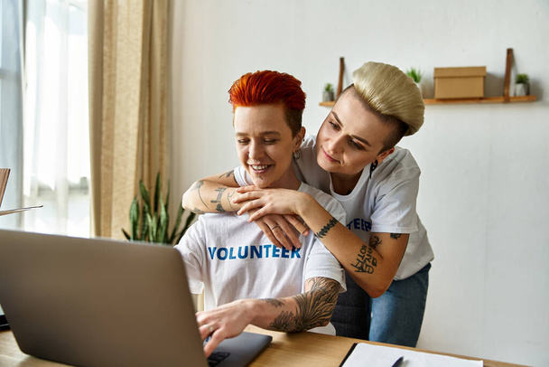 Молодая пара лесбиянок в футболках-волонтерах сидит вместе, работая над ноутбуком для благотворительности. - Фото, изображение