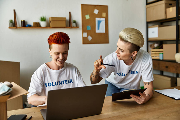 Zwei Frauen in freiwilligen T-Shirts arbeiten gemeinsam an einem Laptop an einem Tisch. - Foto, Bild