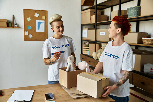 Νεαρό ζευγάρι λεσβιών με εθελοντικά μπλουζάκια στέκονται στο δωμάτιο με κουτιά, ενσαρκώνοντας γενναιοδωρία και συντροφικότητα. - Φωτογραφία, εικόνα