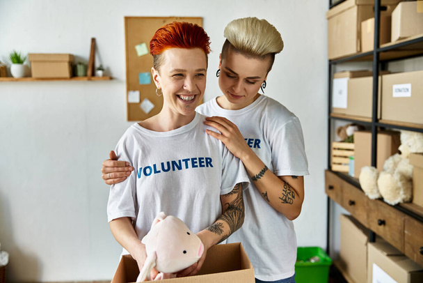 Um jovem e uma mulher, ambos vestindo camisas voluntárias, seguram ternamente um porco de brinquedo aninhado em uma caixa, mostrando amor e cuidado. - Foto, Imagem