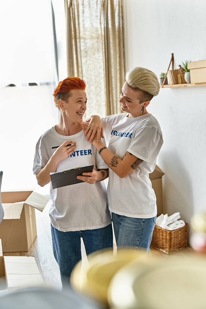 A lesbian couple in volunteer t-shirts, standing side by side, actively engaging in charity work together. - Photo, Image