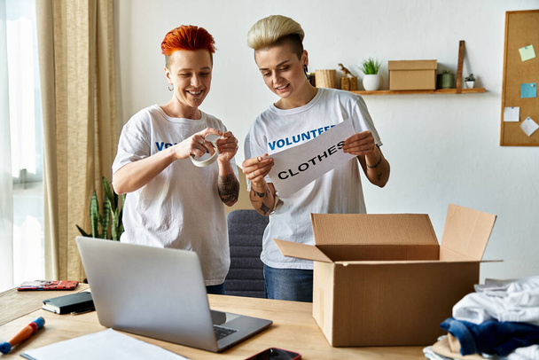 jong lesbisch paar in vrijwilliger t-shirts staan voor een laptop, samen te werken voor een goed doel. - Foto, afbeelding