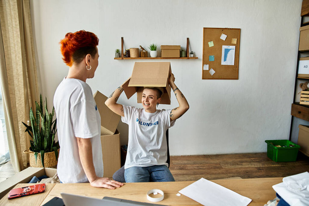 A woman, part of a young lesbian couple, sits on a desk with a box on her head while volunteering in charity work. - Photo, Image