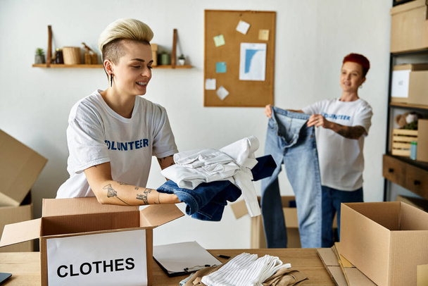 Um jovem casal lésbico, vestindo camisetas voluntárias, descompacta apaixonadamente roupas juntas em um quarto. - Foto, Imagem