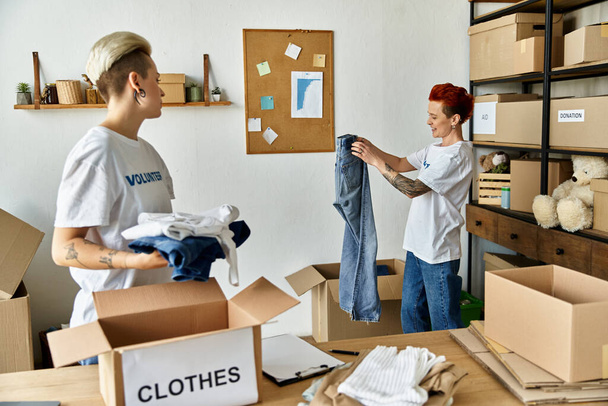 Ein junges lesbisches Paar in freiwilligen T-Shirts packt in einem Raum gemeinsam Kleidung aus, ein herzerwärmender Moment der Teamarbeit und Einheit. - Foto, Bild