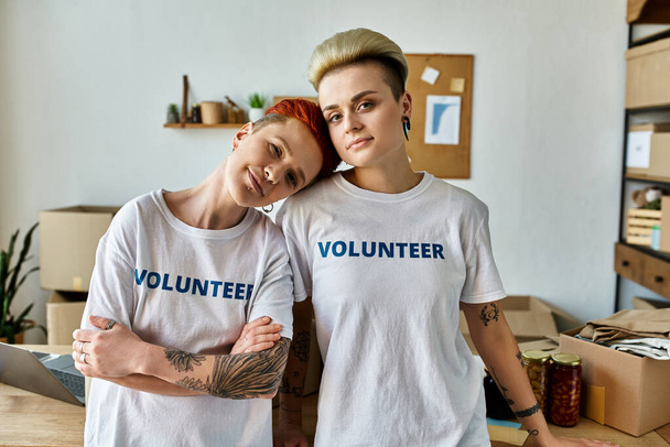 Dwie młode kobiety, noszące ochotnicze koszulki, stoją obok siebie w pokoju, emanując jednością i oddaniem pracy charytatywnej. - Zdjęcie, obraz
