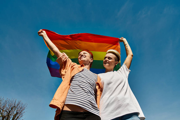 Ένα νεαρό λεσβιακό ζευγάρι που κρατά με χαρά μια σημαία ουράνιου τόξου σε εξωτερικούς χώρους, εκφράζοντας αγάπη και υπερηφάνεια για την ΛΟΑΤΚΙ κοινότητα. - Φωτογραφία, εικόνα