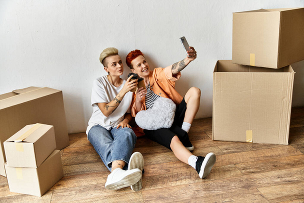 Ein lesbisches Paar sitzt auf dem Boden und knipst gemeinsam ein Selfie in einem Freiwilligenzentrum, umgeben von Kartons für wohltätige Zwecke.. - Foto, Bild
