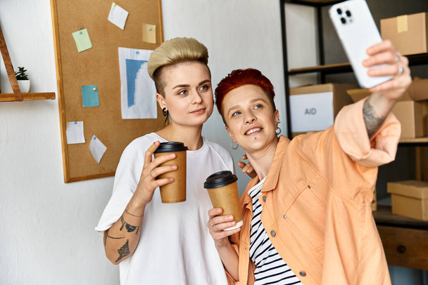 Duas mulheres jovens, um casal lésbico, posam alegremente com uma xícara de café nas mãos enquanto tomam uma selfie em um centro voluntário.. - Foto, Imagem