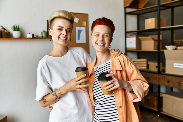 Mladý lesbický pár stojící bok po boku, držící šálky kávy v dobrovolnickém centru obklopeném charitativními krabicemi. - Fotografie, Obrázek